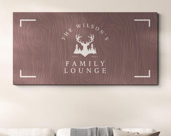 Könnte beinhalten: Ein rechteckiges Holzschild mit dem weißen Text "THE WILSON'S FAMILY LOUNGE", das ein Hirsch- und Baum-Motiv zeigt. Das Schild ist über einem hellgrauen Ecksofa mit dekorativen Kissen und einem Couchtisch aus Holz angebracht.