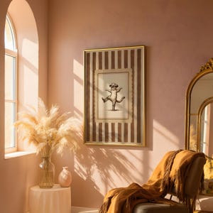 May include: A framed print featuring a whimsical illustration of a cat wearing a hat and dancing. The artwork is set against a striped background in shades of brown and cream, encased in a gold frame. The room has a warm, inviting ambiance with a chair and a vase of pampas grass.