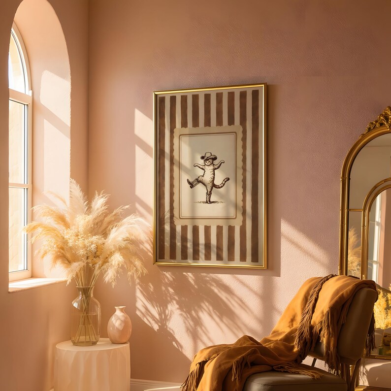 May include: A framed print featuring a whimsical illustration of a cat wearing a hat and dancing. The artwork is set against a striped background in shades of brown and cream, encased in a gold frame. The room has a warm, inviting ambiance with a chair and a vase of pampas grass.