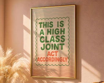May include: A framed art print with the text "THIS IS A HIGH CLASS JOINT ACT ACCORDINGLY" in green and red lettering. The print has a gold frame and is displayed on a pink wall. Decorative pampas grass is visible in the lower left corner.