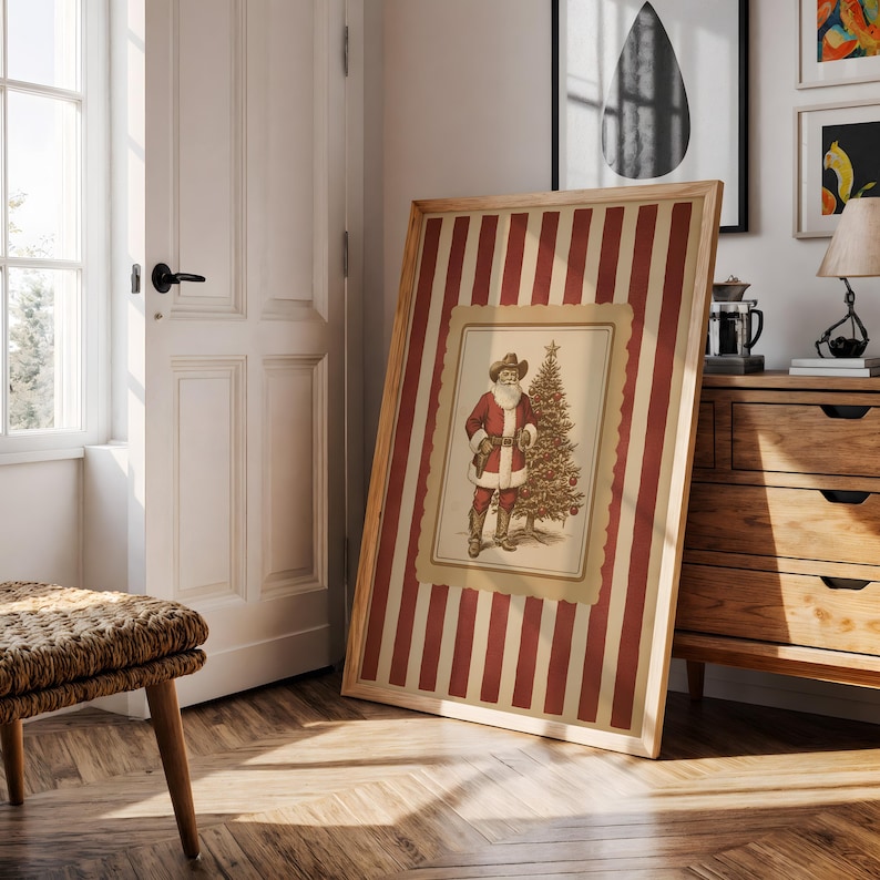 May include: Framed vintage print featuring a cowboy Santa Claus with a Christmas tree. The artwork is set against a red and cream striped background, encased in a wooden frame. The print is leaning against a wall in a room with wooden floors.
