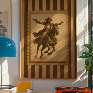 May include: Framed sepia-toned print of a cowboy on horseback, arms outstretched, holding two pistols. The artwork is set against a striped background and framed in a wooden border. The image evokes a vintage Western theme.