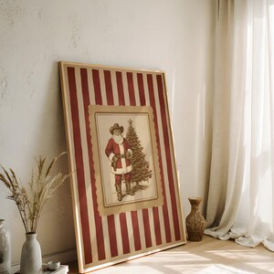 May include: A framed Christmas print featuring a vintage illustration of Santa Claus in a cowboy hat and a Christmas tree. The frame has a red and cream striped pattern. The artwork is leaning against a white wall.
