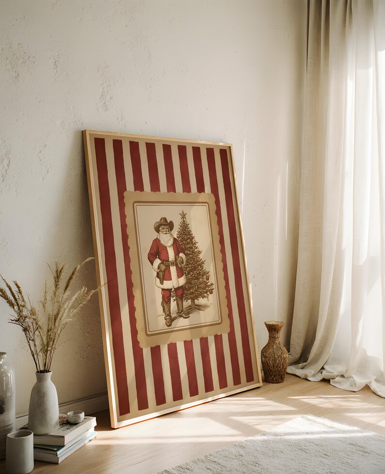 May include: A framed Christmas print featuring a vintage illustration of Santa Claus in a cowboy hat and a Christmas tree. The frame has a red and cream striped pattern. The artwork is leaning against a white wall.