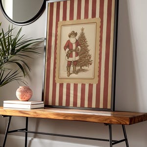 May include: Framed vintage-style Christmas print featuring Santa Claus in a cowboy hat and red coat, standing next to a decorated Christmas tree. The background has red and cream vertical stripes. The print is displayed on a wooden bench.