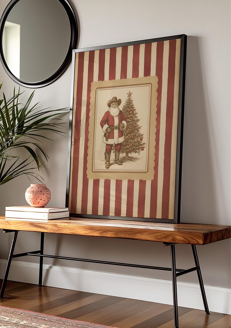 May include: Framed vintage-style Christmas print featuring Santa Claus in a cowboy hat and red coat, standing next to a decorated Christmas tree. The background has red and cream vertical stripes. The print is displayed on a wooden bench.