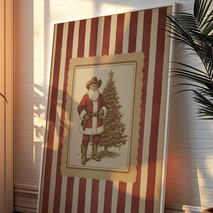 May include: Framed print featuring a cowboy Santa Claus illustration. The artwork depicts Santa in a red suit and cowboy hat, standing next to a decorated Christmas tree. The frame has a striped red and beige pattern, suitable for holiday decor.