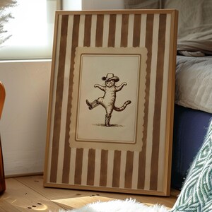 May include: A framed print featuring a whimsical illustration of a cat wearing a cowboy hat and boots, striking a dancing pose. The frame has a light wood finish and a striped background in shades of brown and cream.