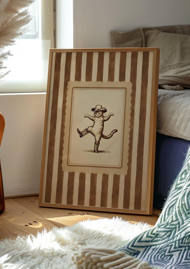 May include: A framed print featuring a whimsical illustration of a cat wearing a cowboy hat and boots, striking a dancing pose. The frame has a light wood finish and a striped background in shades of brown and cream.