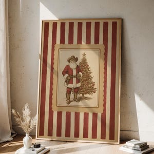 May include: Framed print featuring a cowboy Santa Claus standing next to a Christmas tree. The print has a red and cream striped background. The Santa illustration is in a cream-colored frame with a scalloped edge.