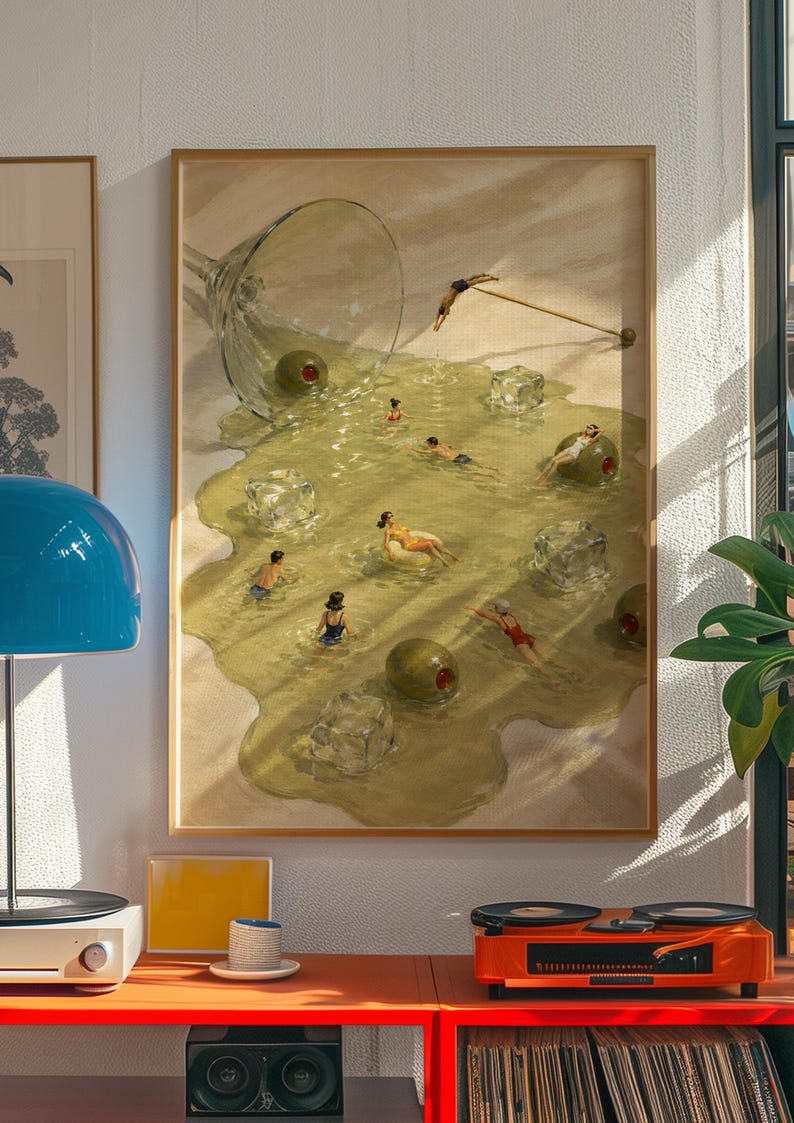 May include: A framed wall art print depicting a spilled martini glass on a sandy surface, with people swimming in the puddle of liquid alongside large green olives and translucent ice cubes, while one person dives from a cocktail pick.