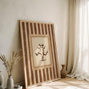 May include: A framed print featuring a sepia-toned illustration of a cat wearing a cowboy hat and boots, striking a dancing pose. The frame has a striped pattern in shades of brown and beige, leaning against a white wall.