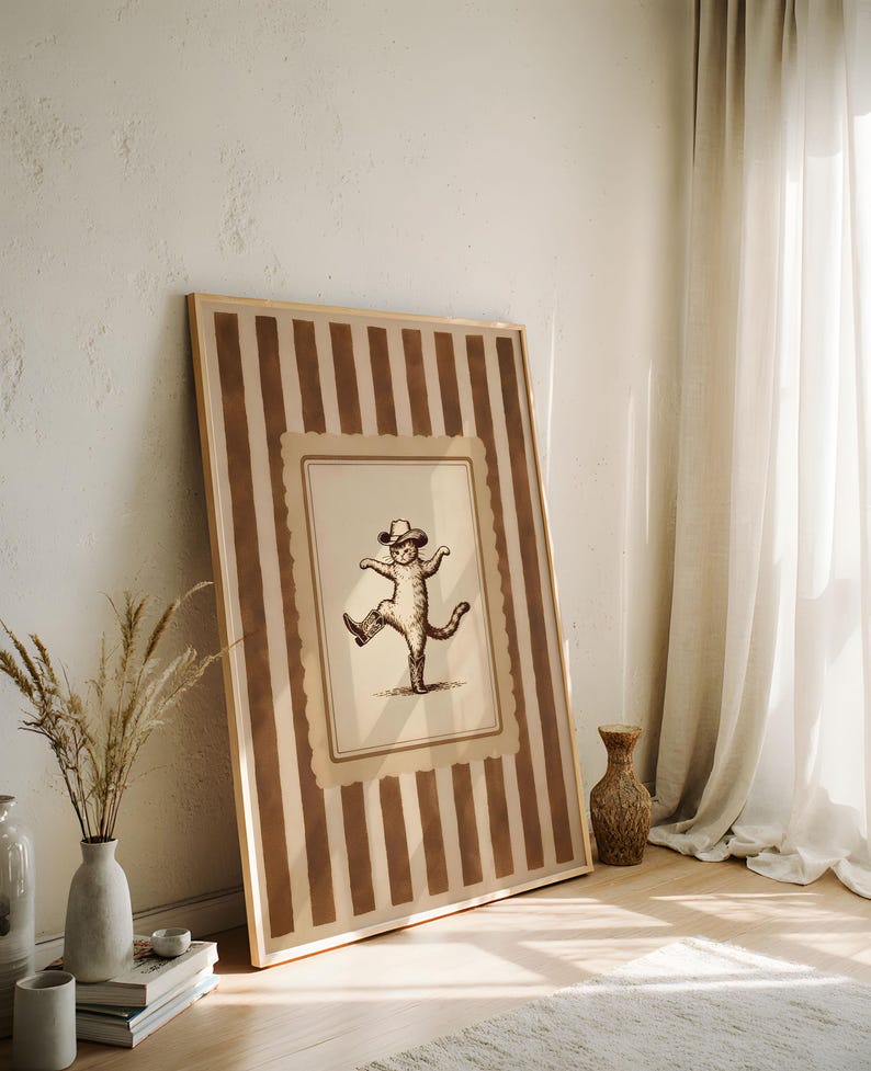May include: A framed print featuring a sepia-toned illustration of a cat wearing a cowboy hat and boots, striking a dancing pose. The frame has a striped pattern in shades of brown and beige, leaning against a white wall.