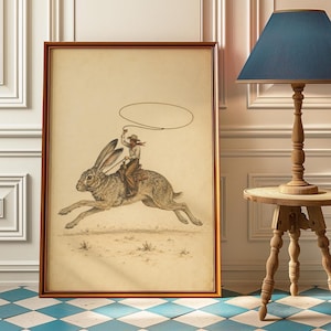 May include: A framed wall art print featuring a detailed illustration of a cowboy riding a large jackrabbit while swinging a lasso. The artwork is rendered in muted earth tones on a cream background, enclosed in a simple brown wooden frame.