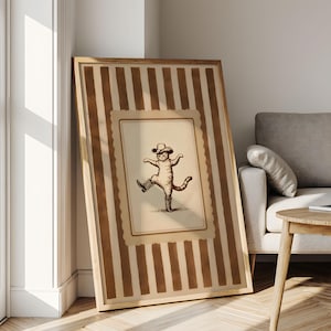 May include: A framed print featuring a whimsical illustration of a cat wearing a cowboy hat and boots, striking a dancing pose. The artwork is set against a cream-colored background with a decorative border, framed by vertical brown stripes.