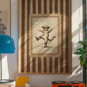 May include: Framed artwork featuring a whimsical illustration of a cat wearing a cowboy hat and boots, striking a dancing pose. The artwork is set against a backdrop of vertical brown stripes and a cream-colored border, creating a vintage aesthetic. The frame is a light brown color.