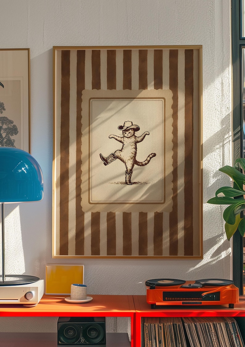 May include: Framed artwork featuring a whimsical illustration of a cat wearing a cowboy hat and boots, striking a dancing pose. The artwork is set against a backdrop of vertical brown stripes and a cream-colored border, creating a vintage aesthetic. The frame is a light brown color.