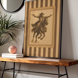 May include: Framed sepia-toned print of a cowboy on horseback, guns drawn, set against a striped background. The artwork is displayed on a wooden bench with black metal legs, complemented by a round mirror and decorative items.