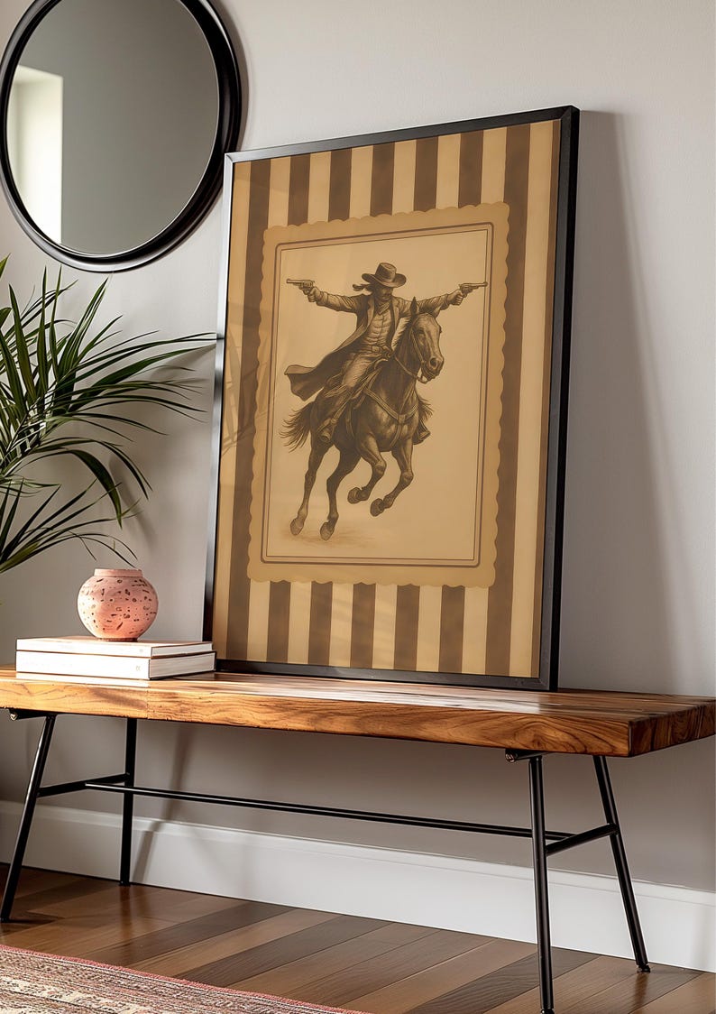 May include: Framed sepia-toned print of a cowboy on horseback, guns drawn, set against a striped background. The artwork is displayed on a wooden bench with black metal legs, complemented by a round mirror and decorative items.