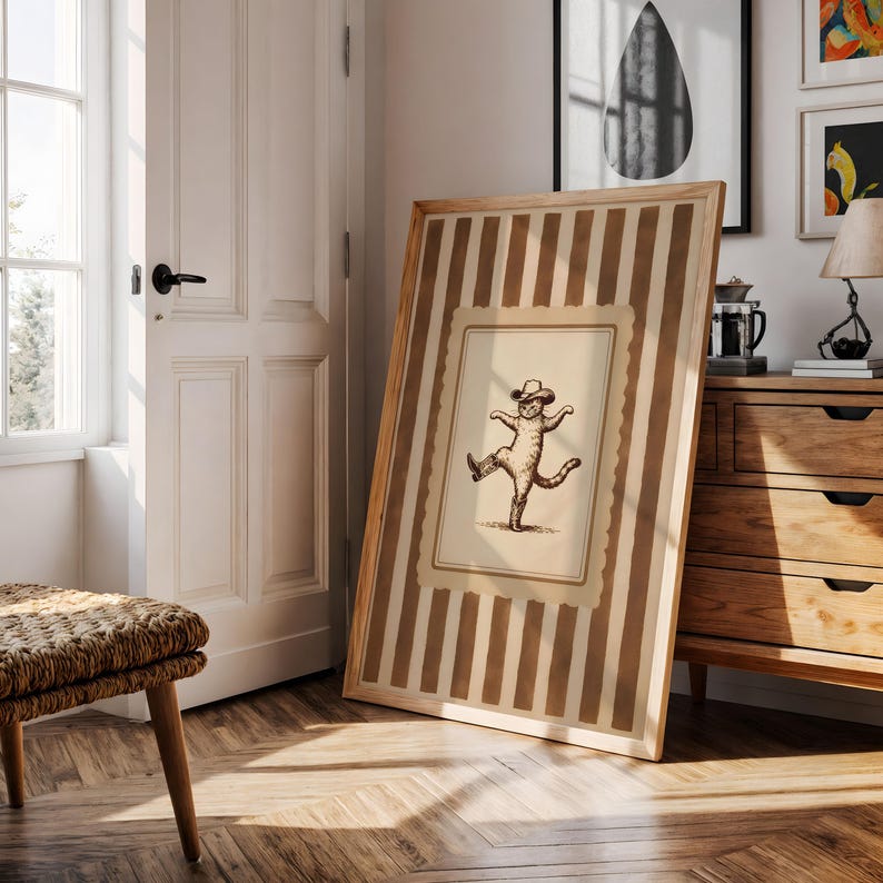 May include: A framed print featuring a whimsical illustration of a cat wearing a hat and boots, striking a dancing pose. The frame has a light wood finish with vertical brown and beige stripes. The artwork is displayed on a wooden floor.