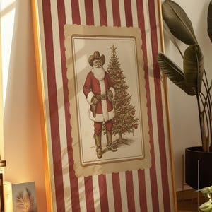 May include: Framed Christmas art featuring a cowboy Santa Claus with a white beard, wearing a cowboy hat and red suit, standing next to a decorated Christmas tree. The background has red and white vertical stripes. The frame is gold.