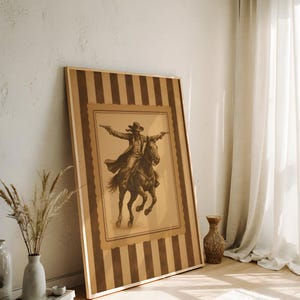 May include: A framed sepia-toned print of a cowboy on horseback, arms outstretched, holding pistols. The artwork is set within a striped border of brown and tan. The frame is leaning against a white wall, next to a vase with dried plants.