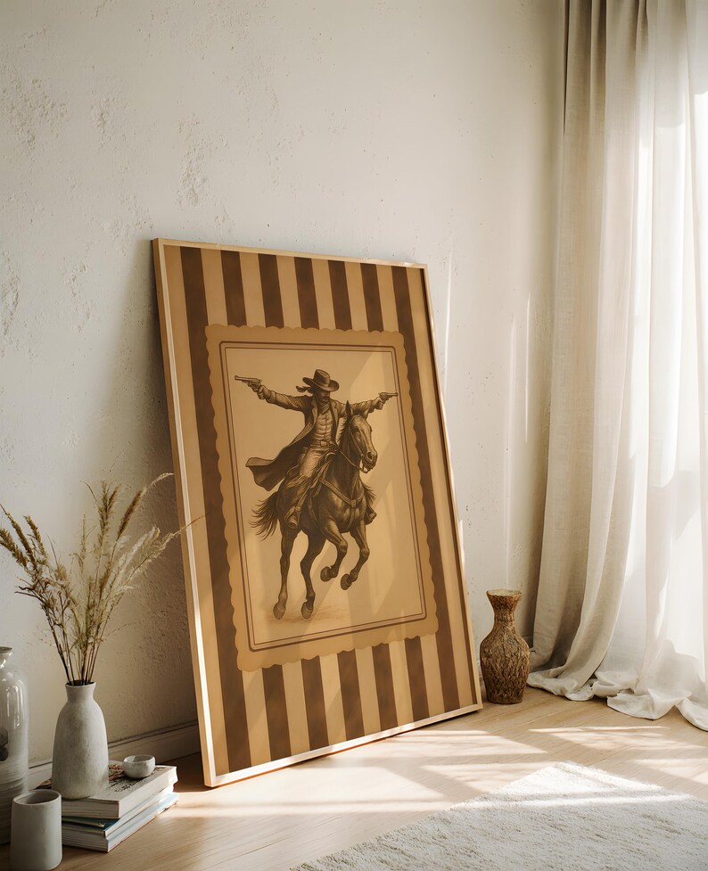 May include: A framed sepia-toned print of a cowboy on horseback, arms outstretched, holding pistols. The artwork is set within a striped border of brown and tan. The frame is leaning against a white wall, next to a vase with dried plants.