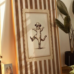 May include: Framed wall art featuring a whimsical illustration of a cat wearing a cowboy hat and boots, striking a dancing pose. The artwork is set against a backdrop of vertical brown and cream stripes, encased in a gold frame.
