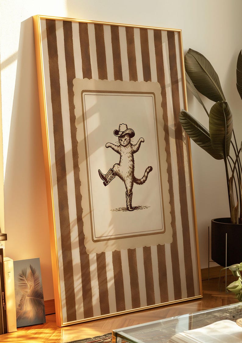 May include: Framed wall art featuring a whimsical illustration of a cat wearing a cowboy hat and boots, striking a dancing pose. The artwork is set against a backdrop of vertical brown and cream stripes, encased in a gold frame.