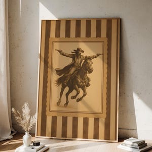 May include: Framed sepia-toned print of a cowboy on horseback, arms outstretched, holding two pistols. The artwork is set against a tan background with a decorative border and a striped frame. The image evokes a classic Western theme.