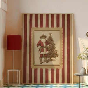 May include: A framed Christmas print featuring Santa Claus in a cowboy hat and attire, standing next to a decorated Christmas tree. The print is set against a red and cream striped background.