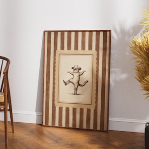 May include: A framed vintage-style print featuring a dancing cat wearing a hat and boots. The artwork is in sepia tones and set within a striped brown and cream frame, leaning against a white wall.