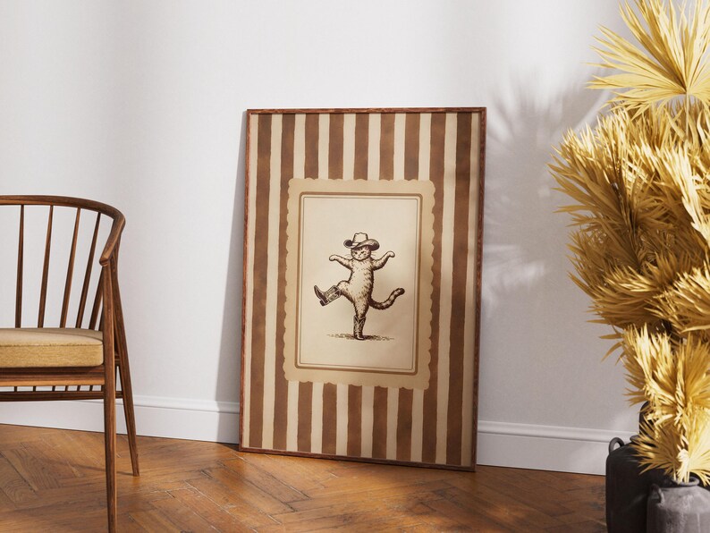 May include: A framed vintage-style print featuring a dancing cat wearing a hat and boots. The artwork is in sepia tones and set within a striped brown and cream frame, leaning against a white wall.