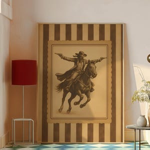 May include: A framed sepia-toned print of a cowboy on horseback, arms outstretched, holding two pistols. The artwork is set against a striped background with a decorative border. The print is displayed in a room with a red lamp and a patterned floor.