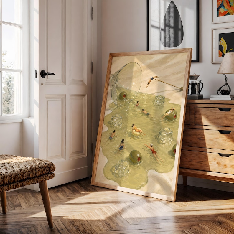 May include: A framed art print featuring a surreal illustration of a spilled martini glass. The liquid forms a pool containing small figures swimming, floating on green olives, and lounging on ice cubes, with a cocktail umbrella resting nearby.