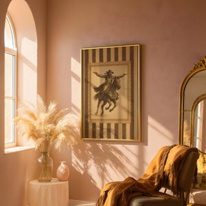 May include: A framed sepia-toned print of a cowboy on horseback, arms outstretched, hangs on a pink wall. The frame has a gold border and a striped background. A gold-framed mirror and a chair with a fringed throw are also in the room.