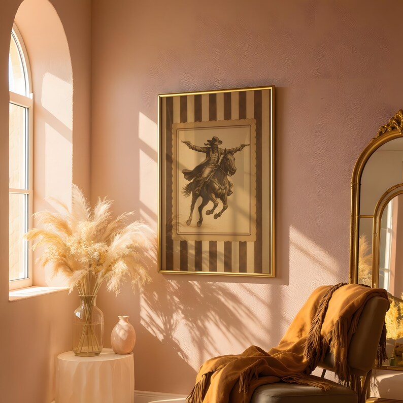 May include: A framed sepia-toned print of a cowboy on horseback, arms outstretched, hangs on a pink wall. The frame has a gold border and a striped background. A gold-framed mirror and a chair with a fringed throw are also in the room.