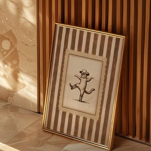 May include: A framed print featuring a whimsical illustration of a cat wearing a cowboy hat and boots, striking a dancing pose. The frame is gold, and the background has vertical brown and white stripes. The print is leaning against a wooden wall.