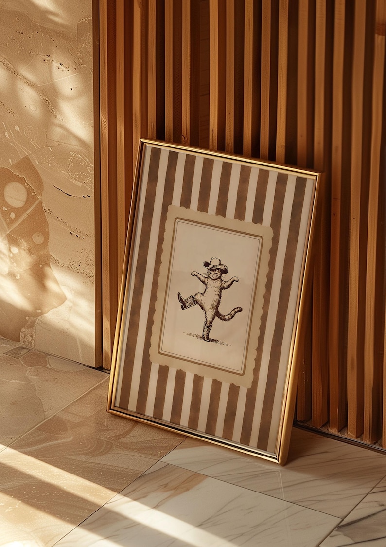 May include: A framed print featuring a whimsical illustration of a cat wearing a cowboy hat and boots, striking a dancing pose. The frame is gold, and the background has vertical brown and white stripes. The print is leaning against a wooden wall.