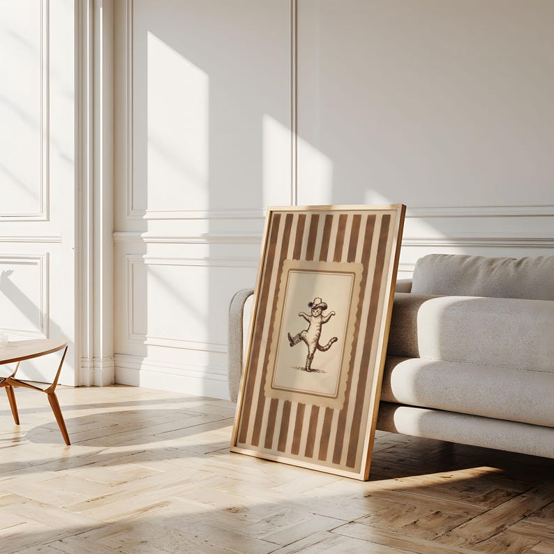 May include: A framed artwork featuring a whimsical illustration of a dancing cat wearing a hat. The artwork is set within a frame with vertical brown stripes and a cream-colored border, leaning against a sofa in a well-lit room.