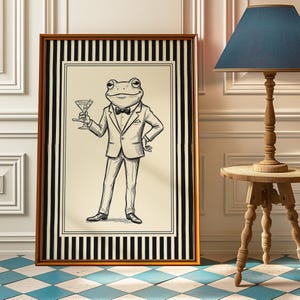 May include: Framed art print featuring a dapper frog in a suit holding a martini. The black and white illustration is set against a cream background, framed with a gold border and black and white stripes. The artwork is displayed on a blue and white checkered floor.
