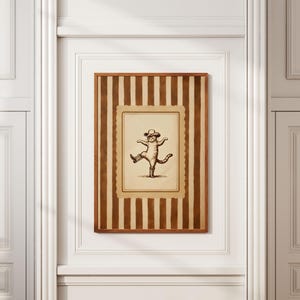 May include: A framed print featuring a whimsical illustration of a cat wearing a hat and dancing. The artwork is set against a striped background in shades of brown and beige, encased in a wooden frame. The print is mounted on a white wall.