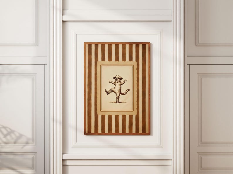 May include: A framed print featuring a whimsical illustration of a cat wearing a hat and dancing. The artwork is set against a striped background in shades of brown and beige, encased in a wooden frame. The print is mounted on a white wall.