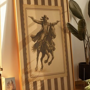 May include: Framed artwork featuring a sepia-toned illustration of a cowboy on horseback, holding two pistols. The artwork is in a gold frame and has a striped background.