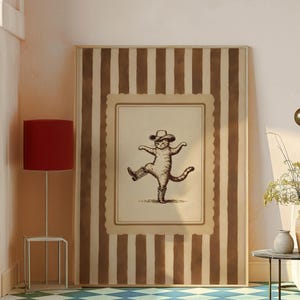 May include: A framed print featuring a whimsical illustration of a cat wearing a cowboy hat and boots, striking a dancing pose. The artwork is set against a backdrop of brown and cream vertical stripes, creating a playful and rustic aesthetic.