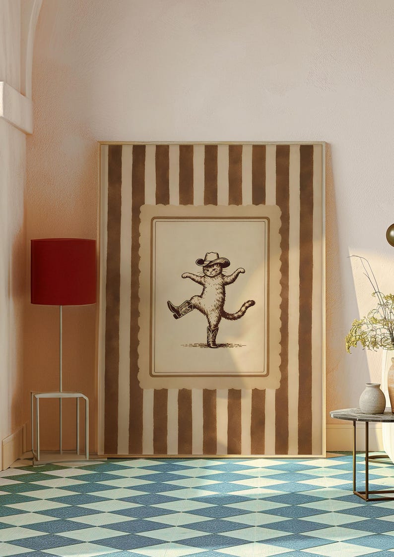 May include: A framed print featuring a whimsical illustration of a cat wearing a cowboy hat and boots, striking a dancing pose. The artwork is set against a backdrop of brown and cream vertical stripes, creating a playful and rustic aesthetic.