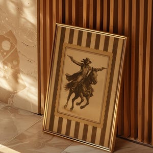 May include: Framed sepia-toned print of a cowboy on horseback, firing two pistols. The artwork is set within a striped border and a gold frame. The print leans against a wooden slatted wall and a tiled floor.