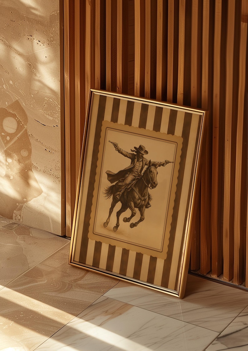 May include: Framed sepia-toned print of a cowboy on horseback, firing two pistols. The artwork is set within a striped border and a gold frame. The print leans against a wooden slatted wall and a tiled floor.