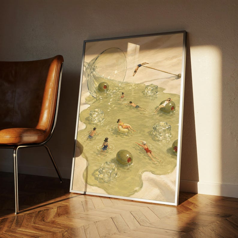 May include: A framed rectangular art print featuring a surreal illustration of a spilled martini glass on a sandy surface. The spilled liquid forms a pool containing swimming figures, large green olives, and translucent ice cubes.