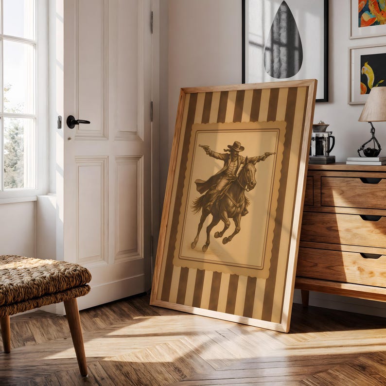 May include: A framed sepia-toned print of a cowboy on horseback, guns drawn, set against a striped background. The artwork is in a light wood frame, leaning against a wall in a room with a wooden floor and a white door.